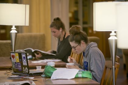 Two students sittin gat a table with a lamp studying. One has a computer the other is reading a book.